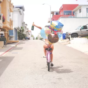 A woman riding a bike, holding a champagne bottle and balloons.